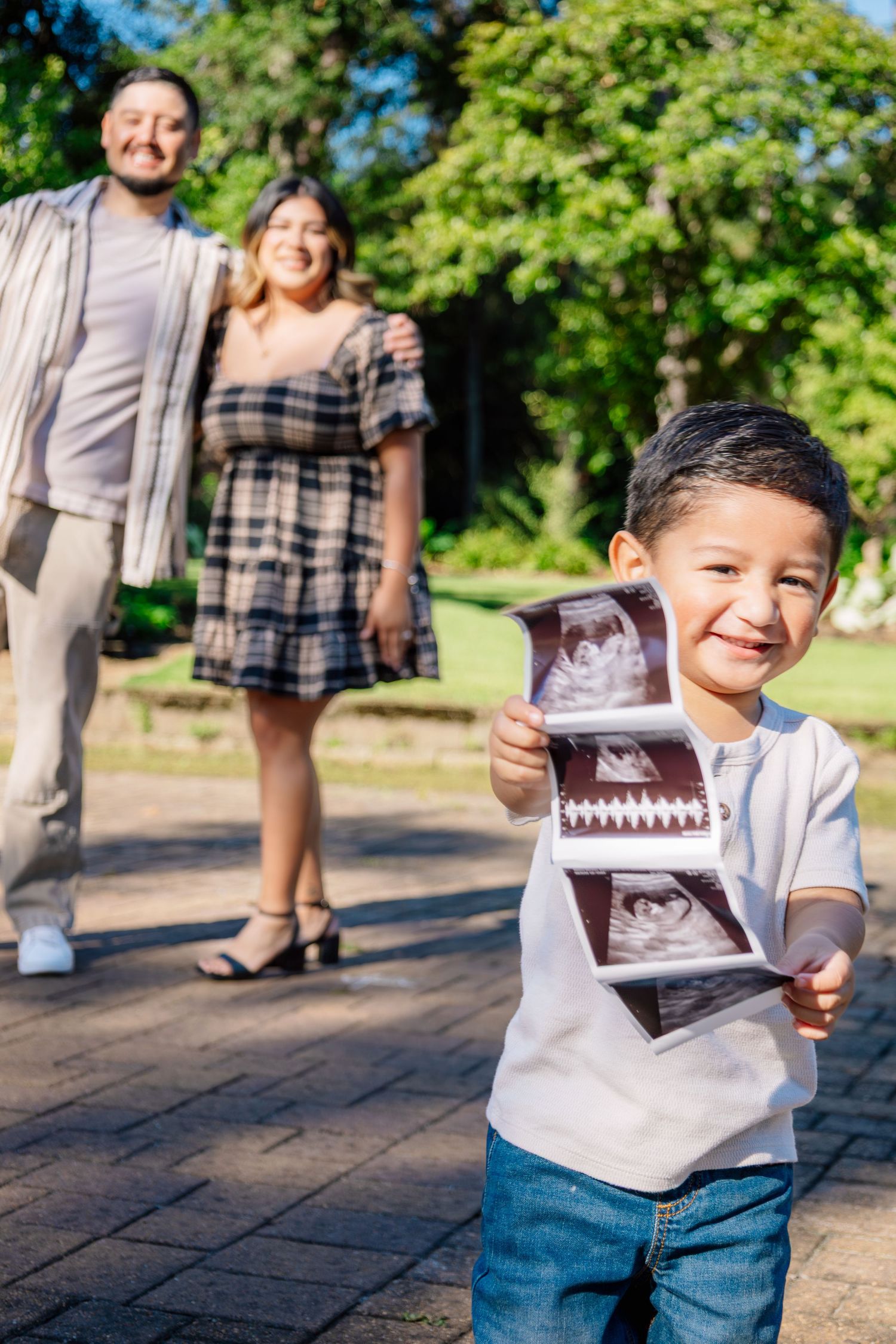 Smiling boy holds ultrasound photos while his parents stand behind him in a sunny park.