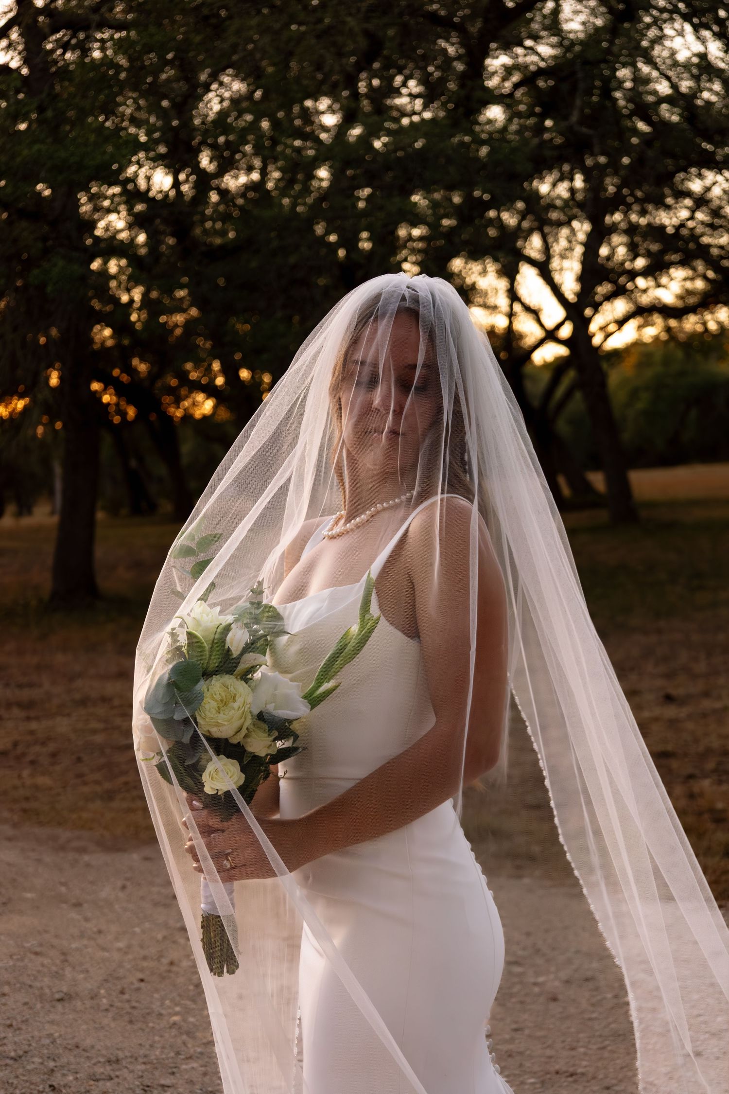 Bride in white gown and veil holding a bouquet of white roses at golden hour sunset outdoors.