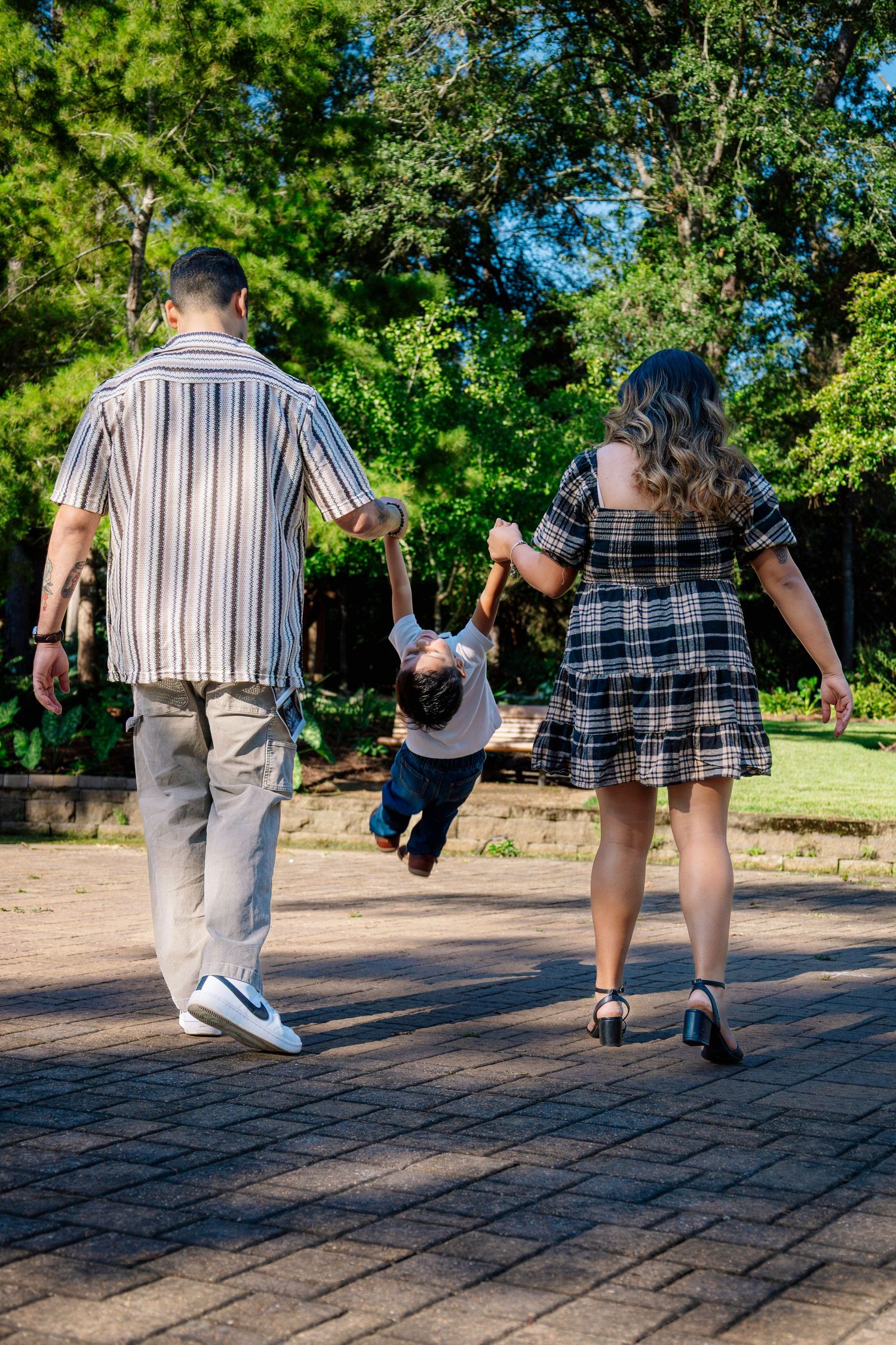 Family of three walking on brick path as parents swing their young child between them in a sunny park.