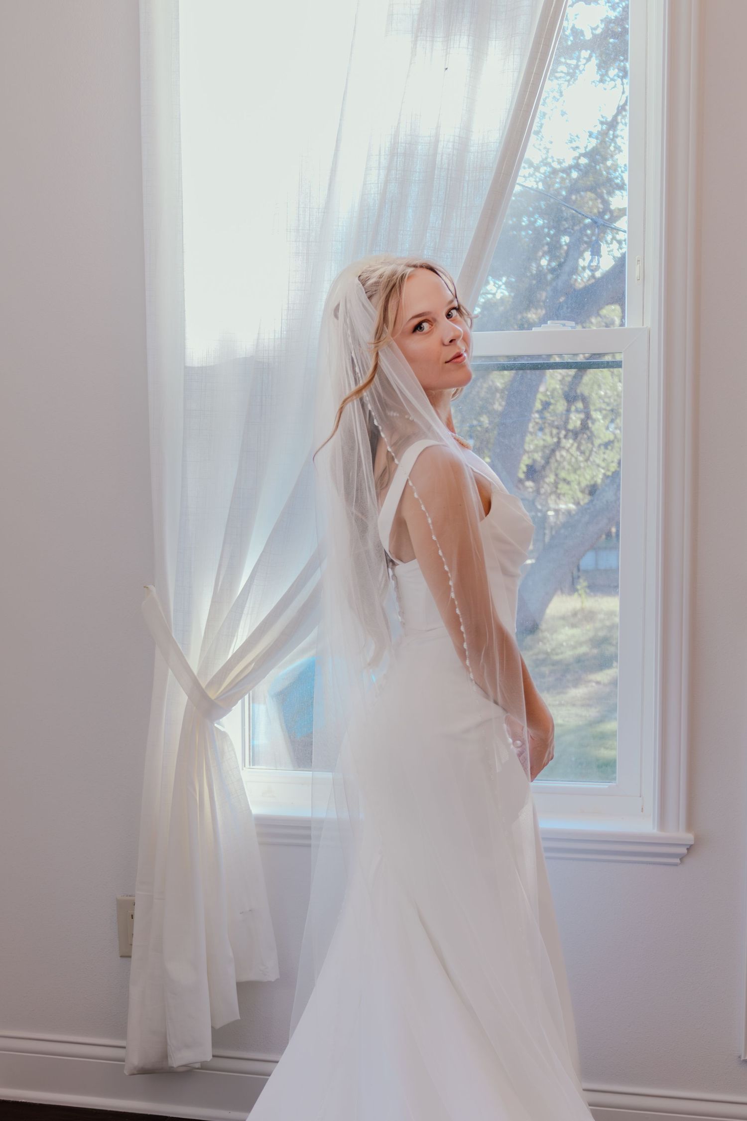 Elegant bride in a white fitted gown and pearl-edged veil posing gracefully by a sunlit window.