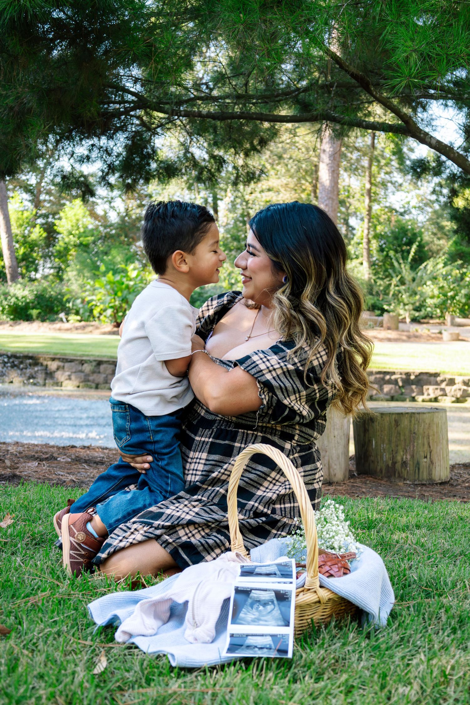 Mother in plaid dress sitting on grass with toddler son, sharing a tender nose-to-nose moment in a park.