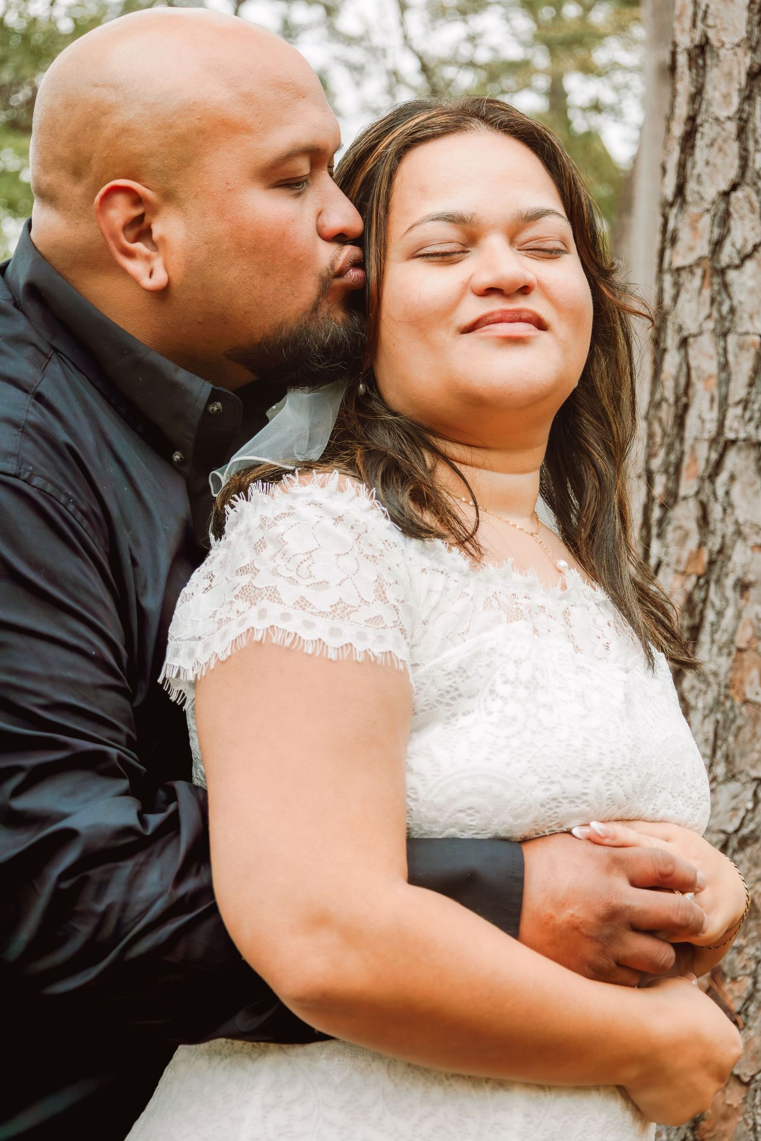 Groom kissing bride on cheek outdoors near tree trunk during wedding photo session.
