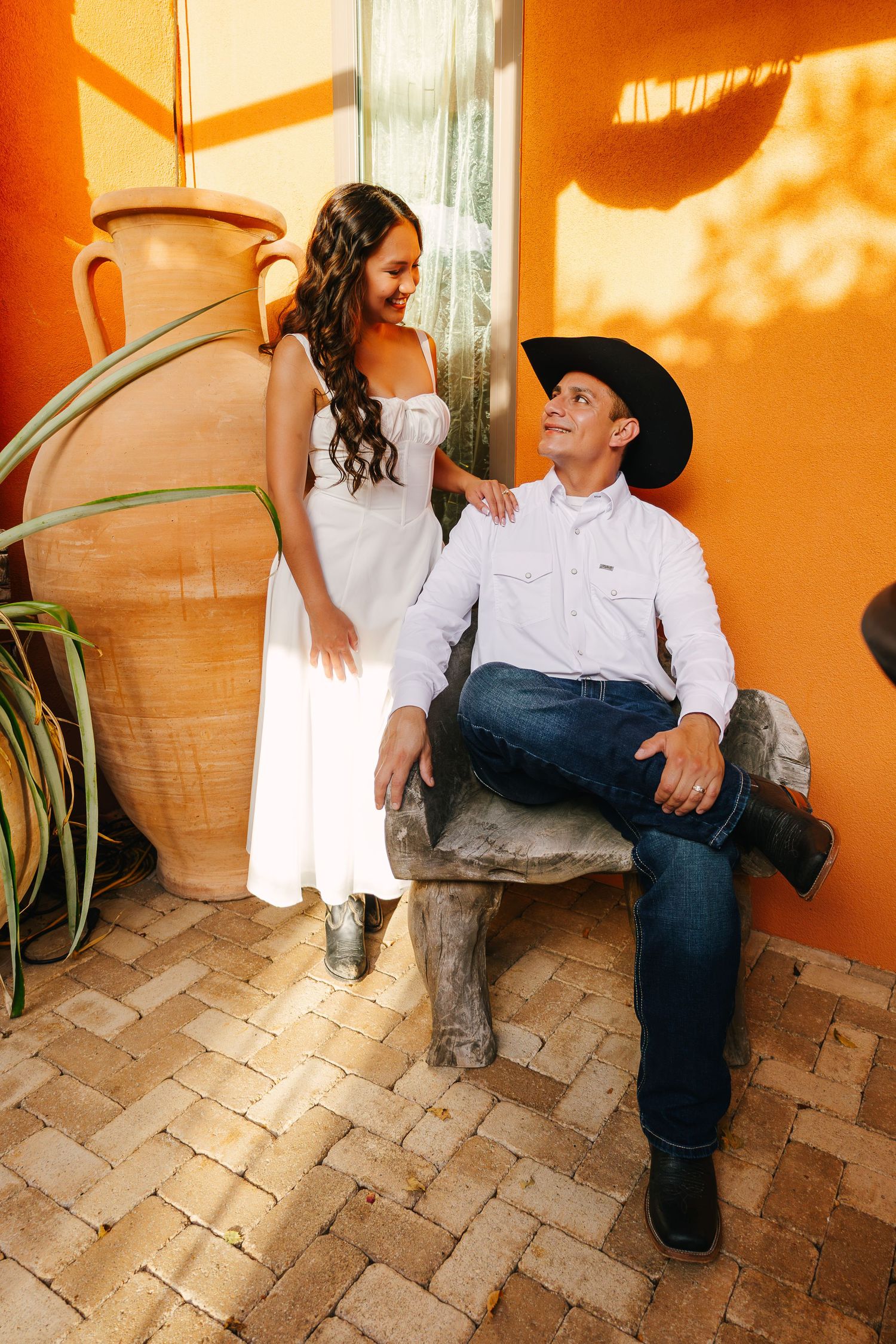 Couple in southwestern setting, woman in white dress and man in cowboy hat sitting on stone chair near large terracotta pot.