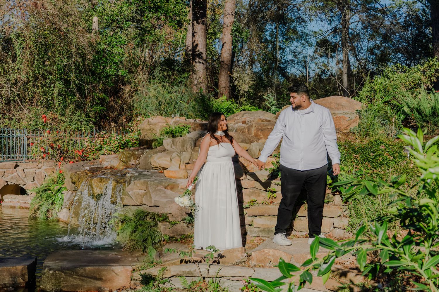 Couple holding hands by a stone waterfall in a lush garden, woman in white dress, man in grey sweater.