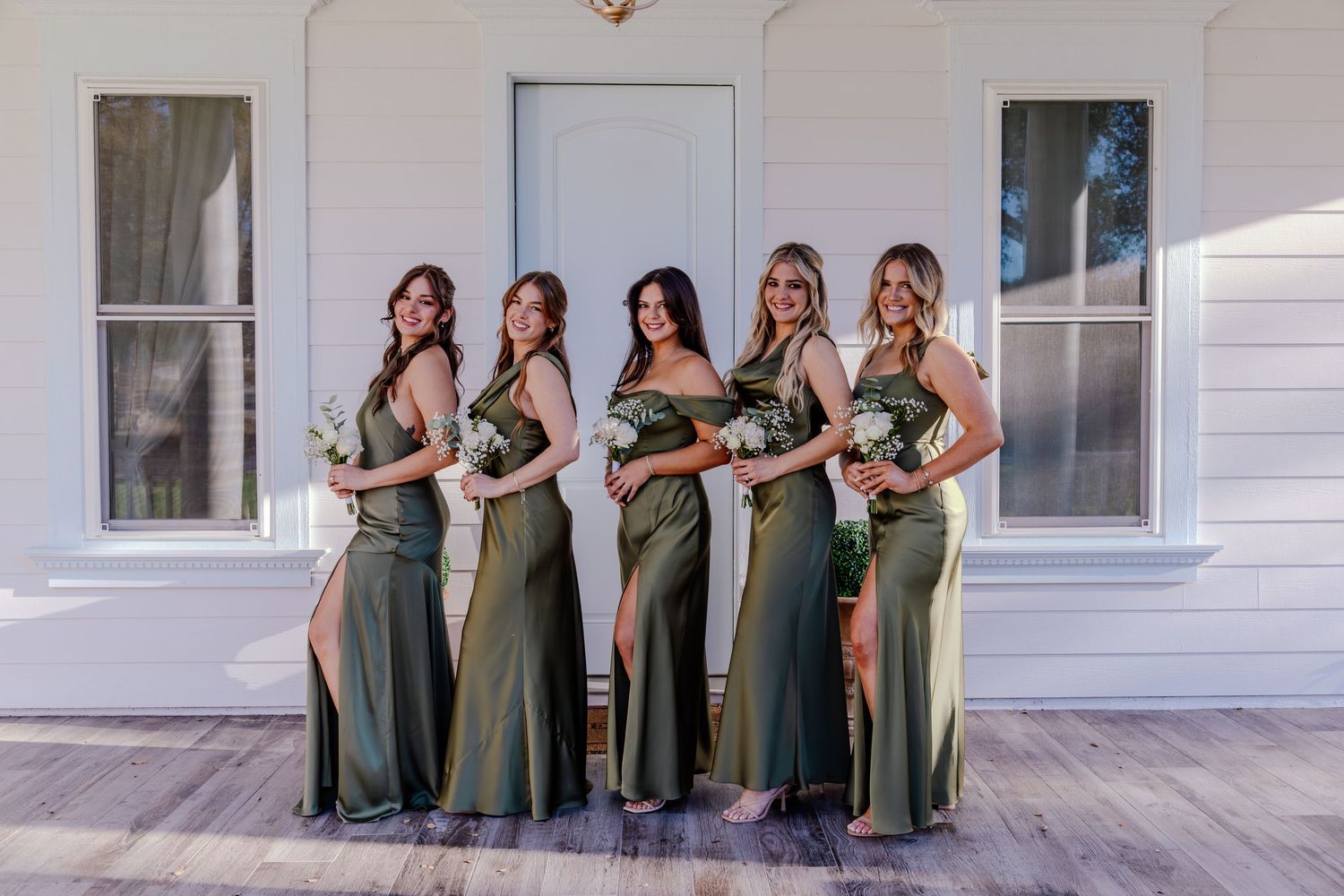Five bridesmaids in olive green dresses posing together in front of a white building.