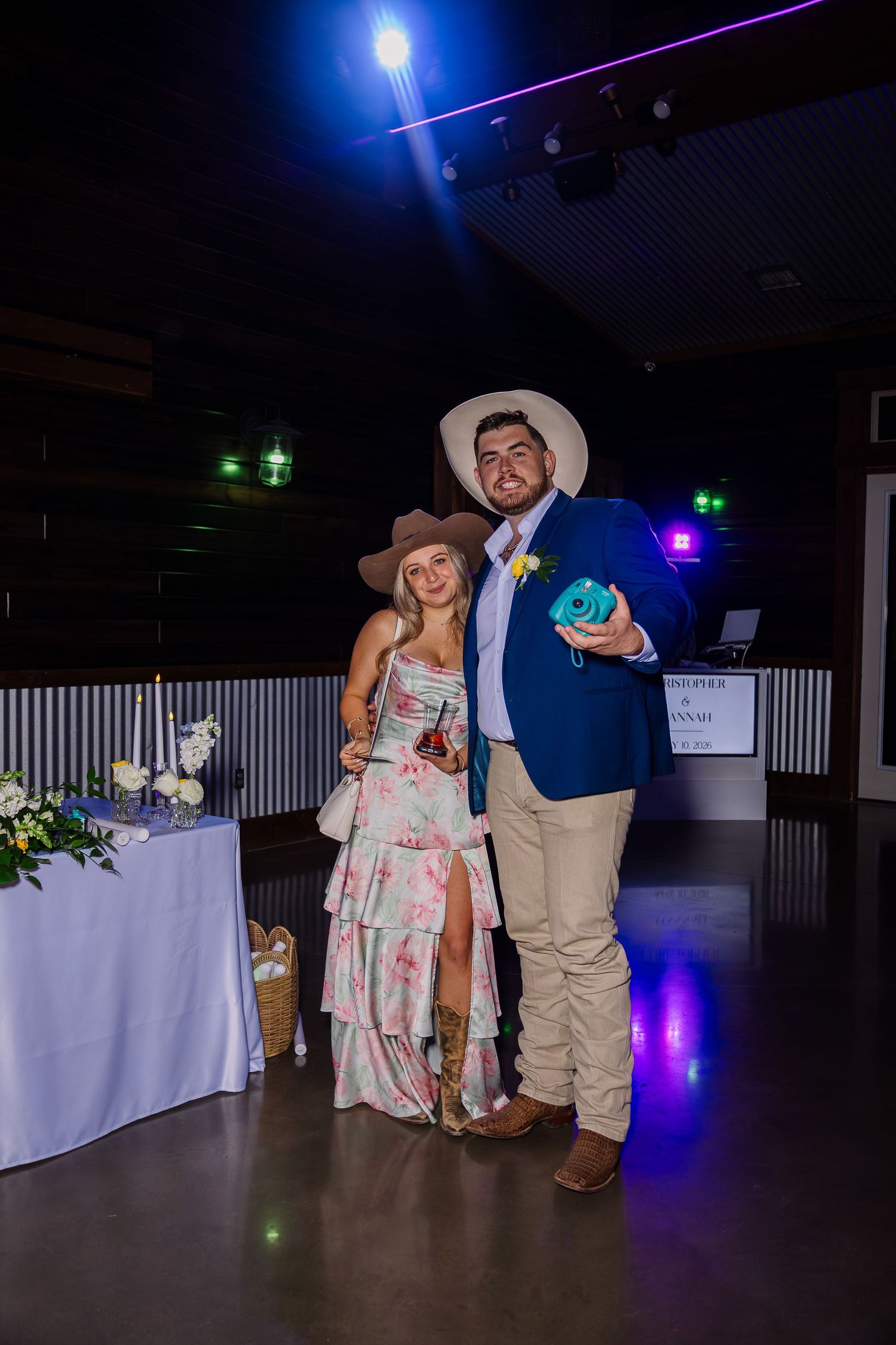 Couple in cowboy hats posing at an indoor event, man in blue blazer holding teal camera, woman in floral dress.