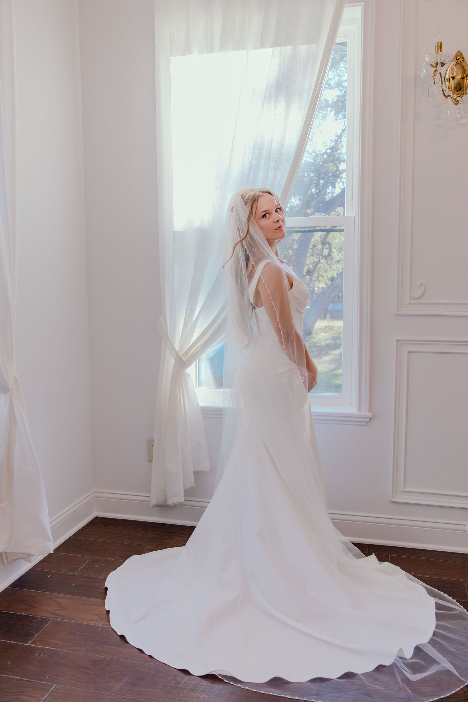 Elegant bride in a white gown and veil standing by a bright window in a white room.