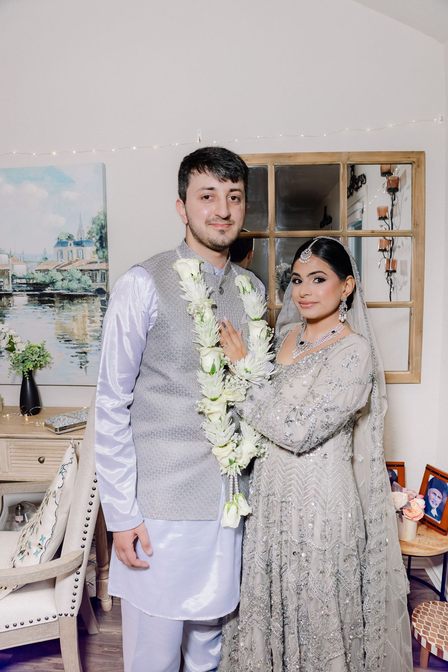 South Asian couple in traditional wedding attire, man wearing floral garland, woman in embroidered gray lehenga, posing indoors.