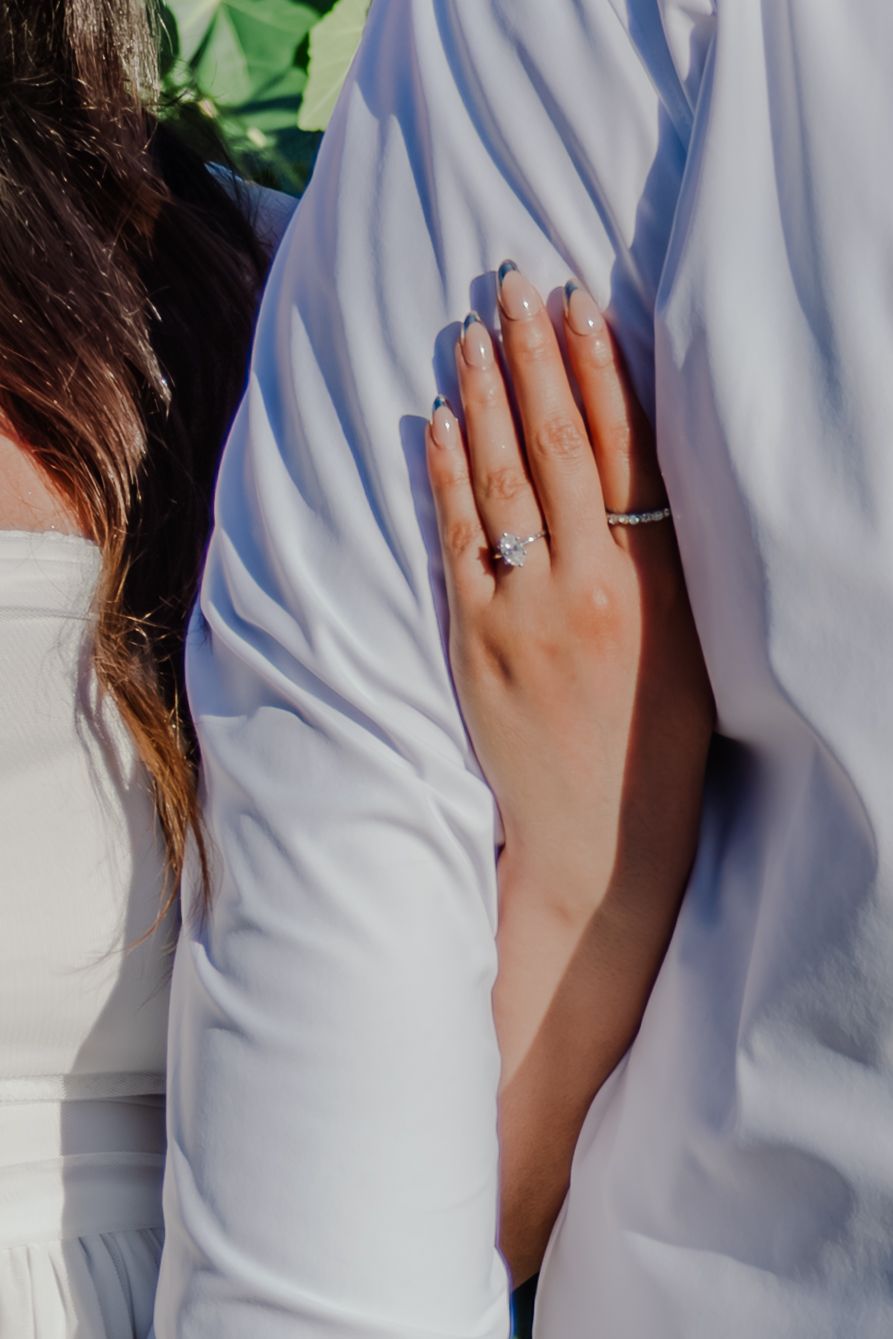 Close-up of bride's hand with diamond engagement ring and wedding band resting on groom's white suit sleeve.