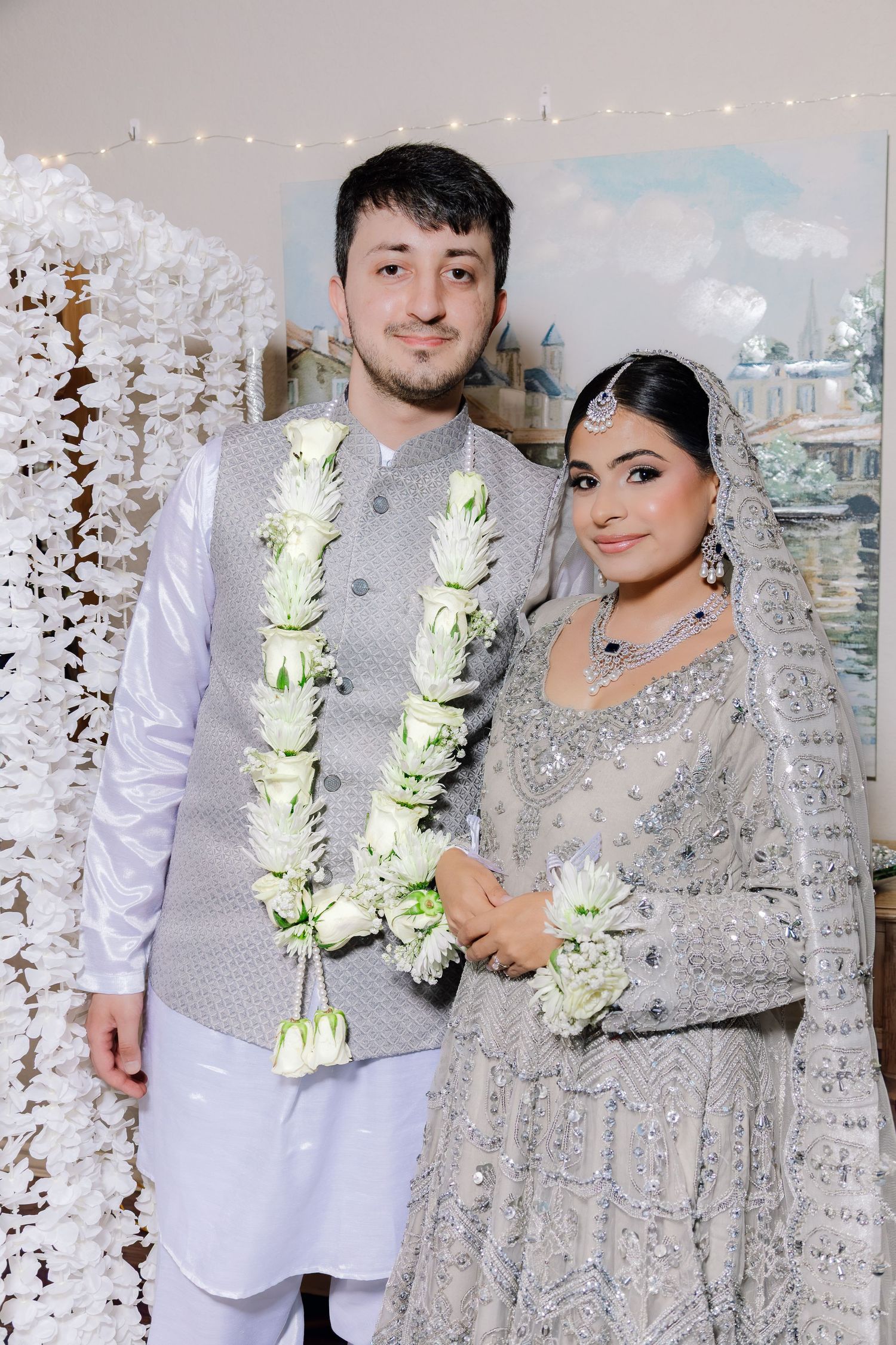 Bride and groom in coordinating grey embroidered outfits with white flower garlands posing at a South Asian wedding.