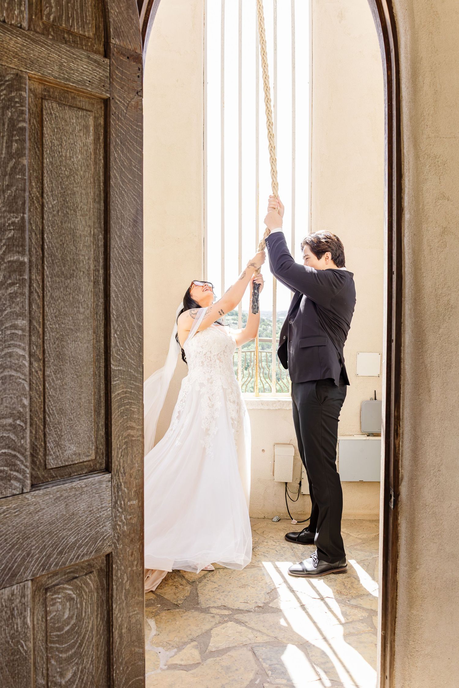 Bride and groom pulling a church bell rope together in a sunlit chapel doorway on their wedding day.