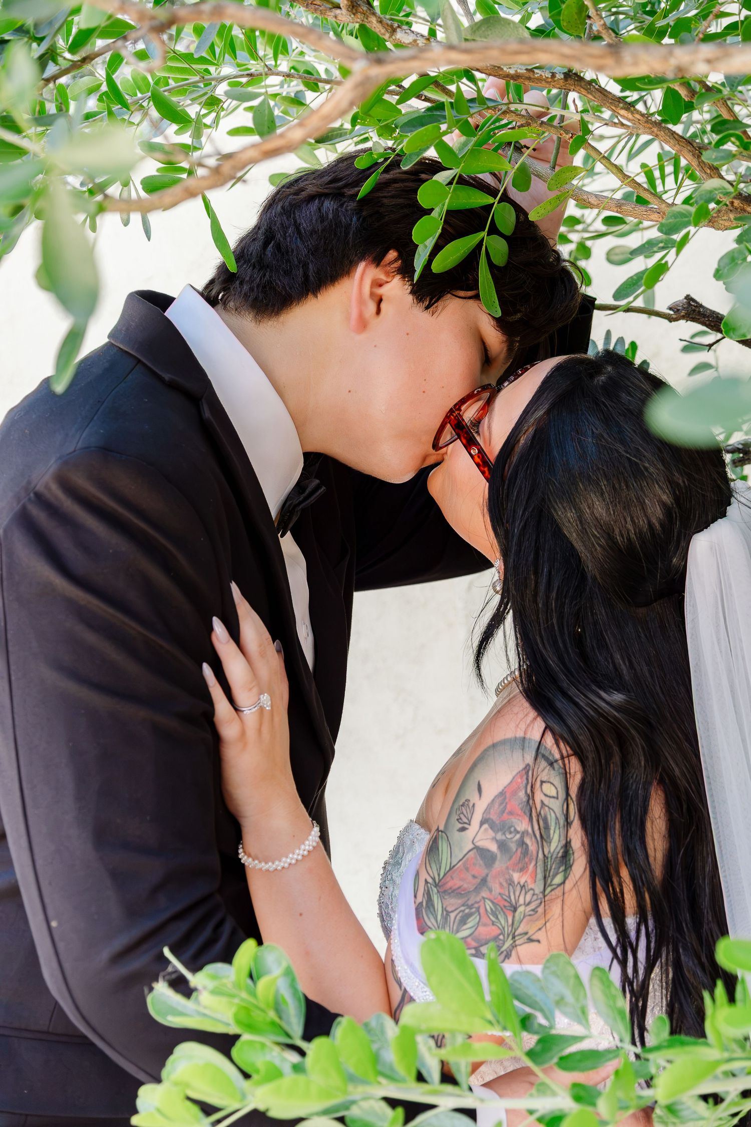A couple shares a romantic kiss under green tree branches, bride with tattoos and veil, groom in black suit.