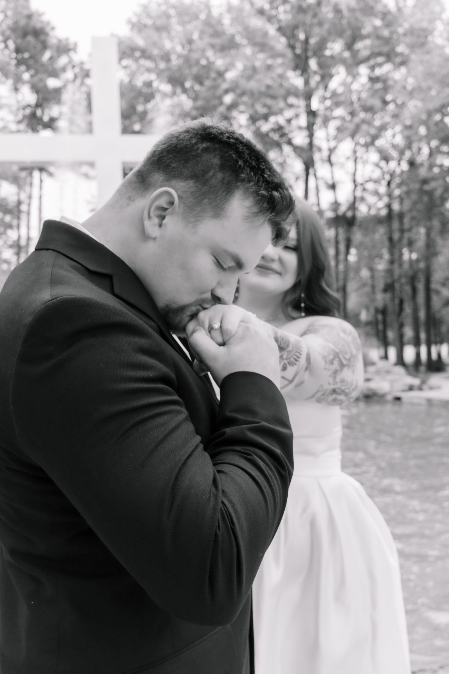 Black and white photo of groom kissing bride's hand near a white cross with trees in background.