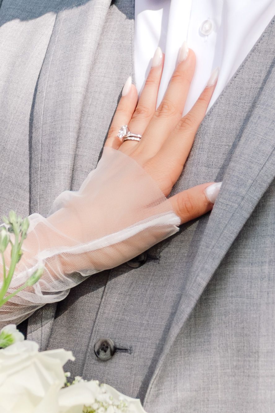 Bride's hand with diamond engagement ring and wedding band resting on groom's gray suit jacket.