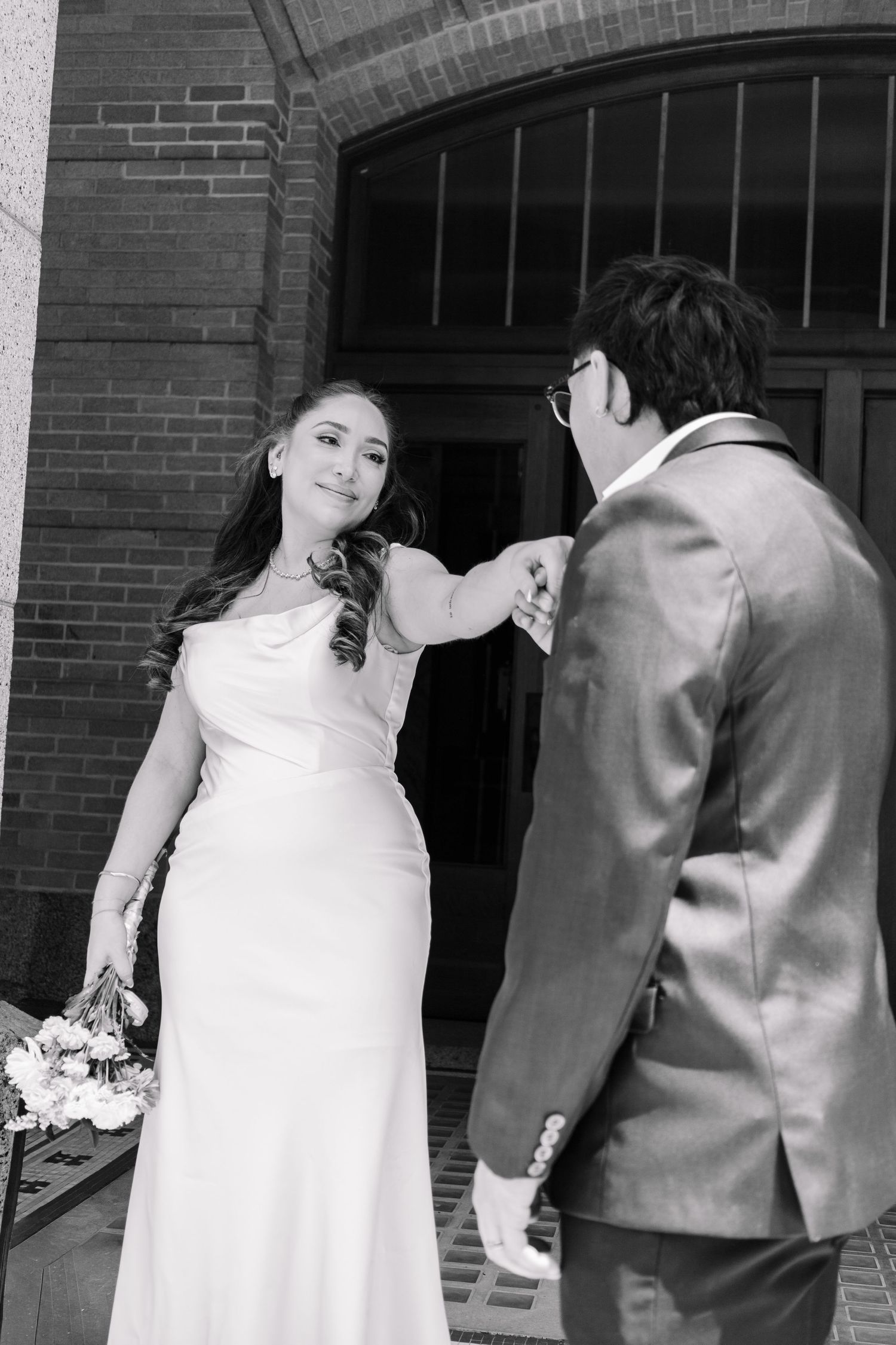 Black and white photo of bride extending hand to groom outside a brick building.