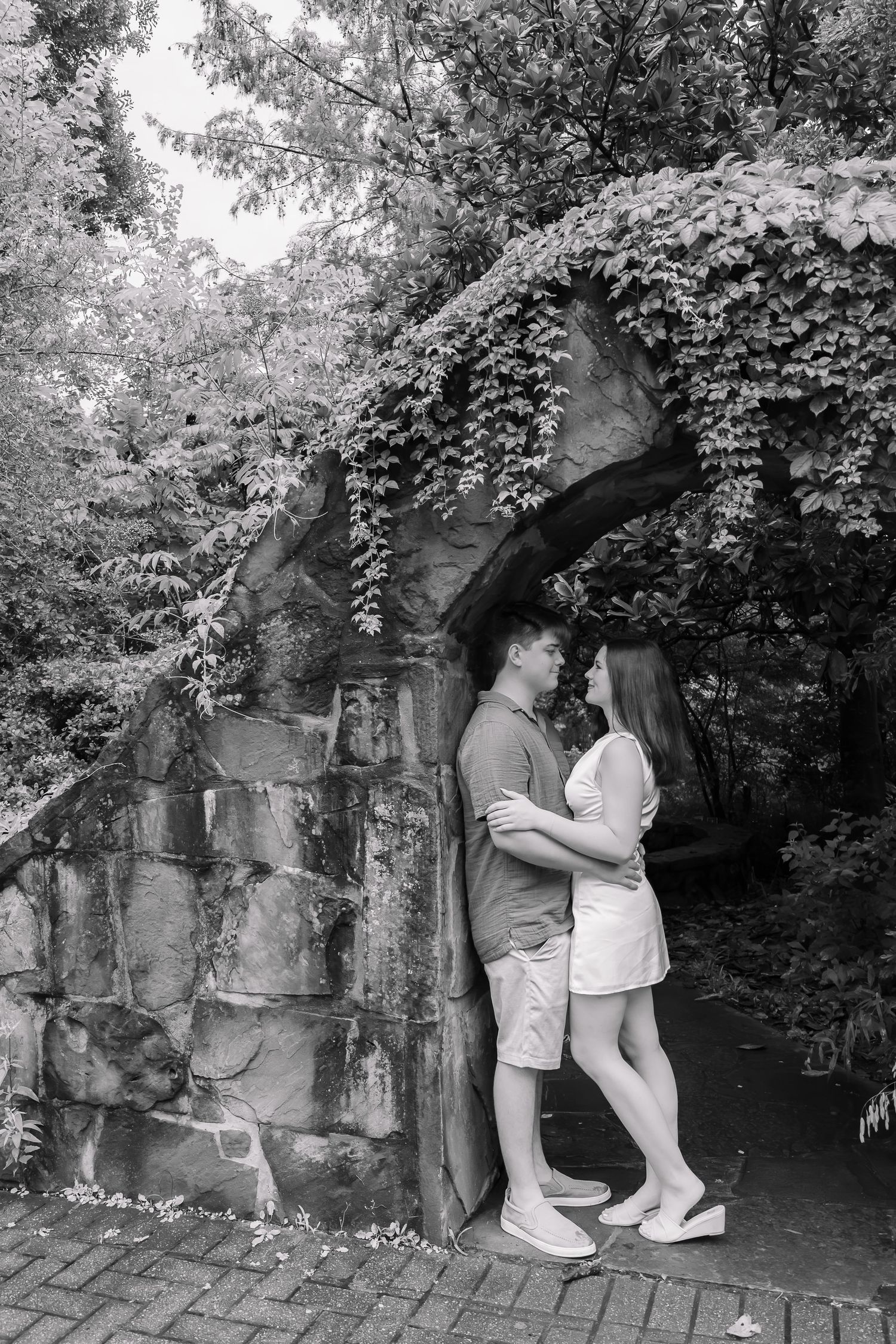 Black and white photo of a romantic couple embracing under a stone arch covered in ivy in a lush garden.