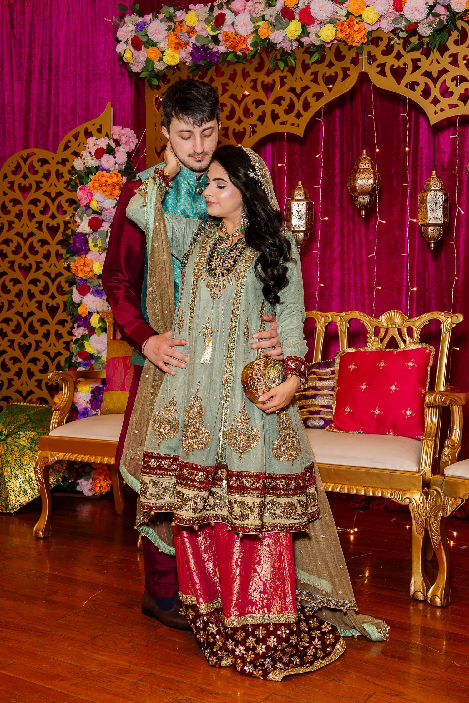 Couple in traditional South Asian wedding attire posing together at an ornate decorated venue with floral backdrop.