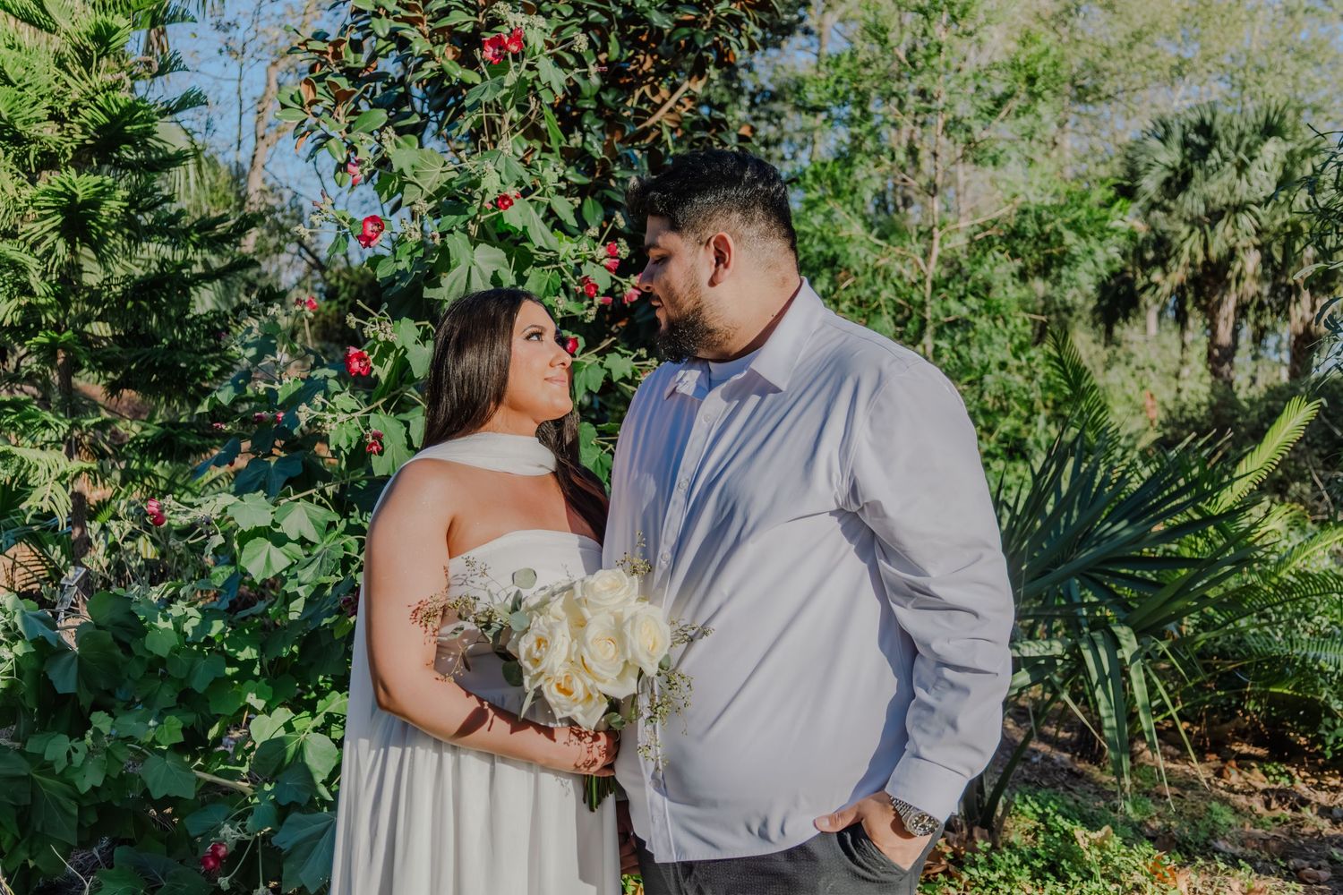 Couple in wedding attire gazing at each other surrounded by lush green garden foliage.