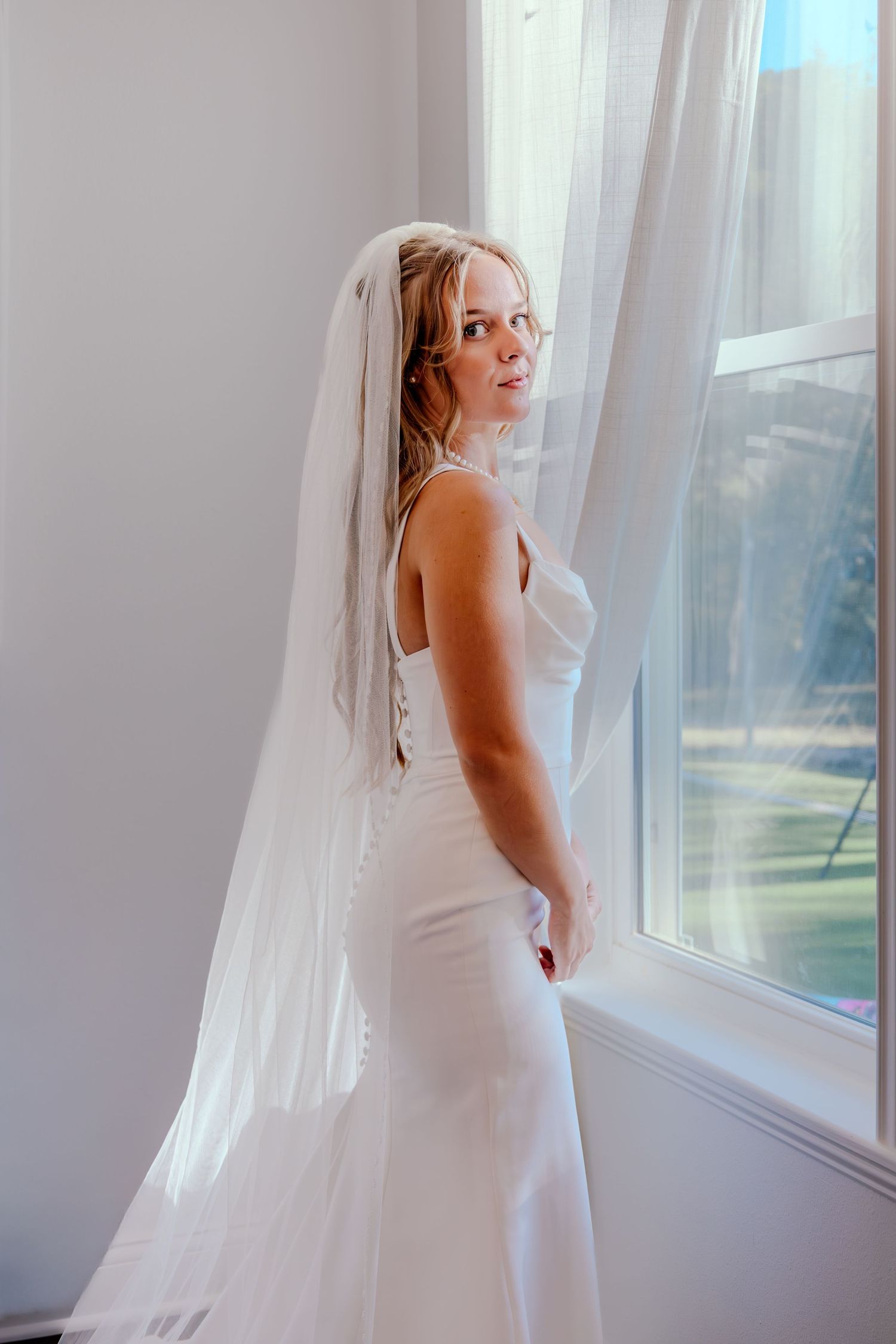 Bride in a fitted white gown and veil gazing over her shoulder by a bright window.