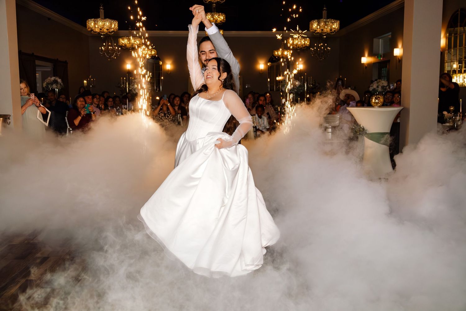 Bride and groom share first dance surrounded by fog machines and sparklers at elegant wedding reception.