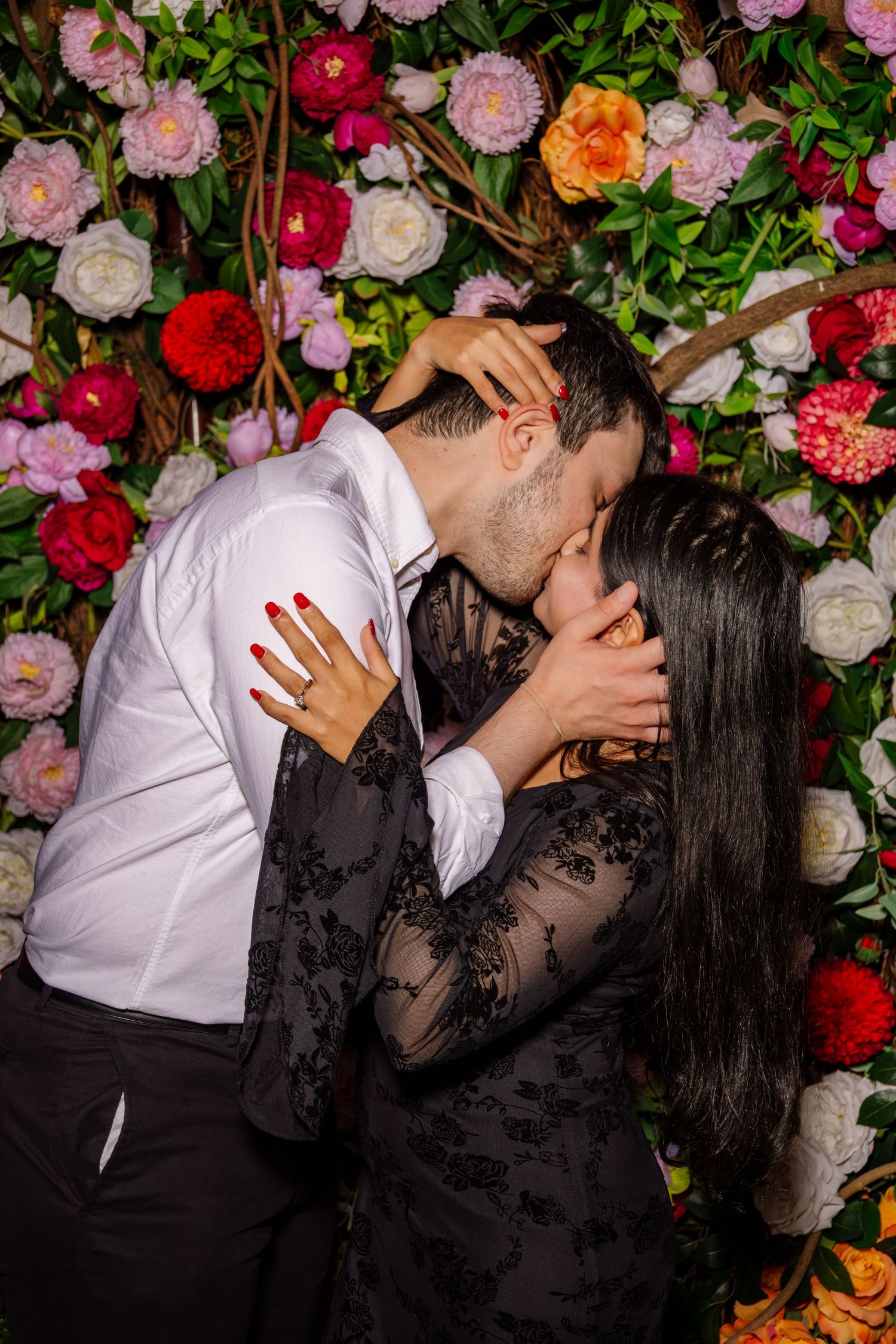 Couple shares a passionate kiss in front of a vibrant colorful floral wall backdrop at their engagement.