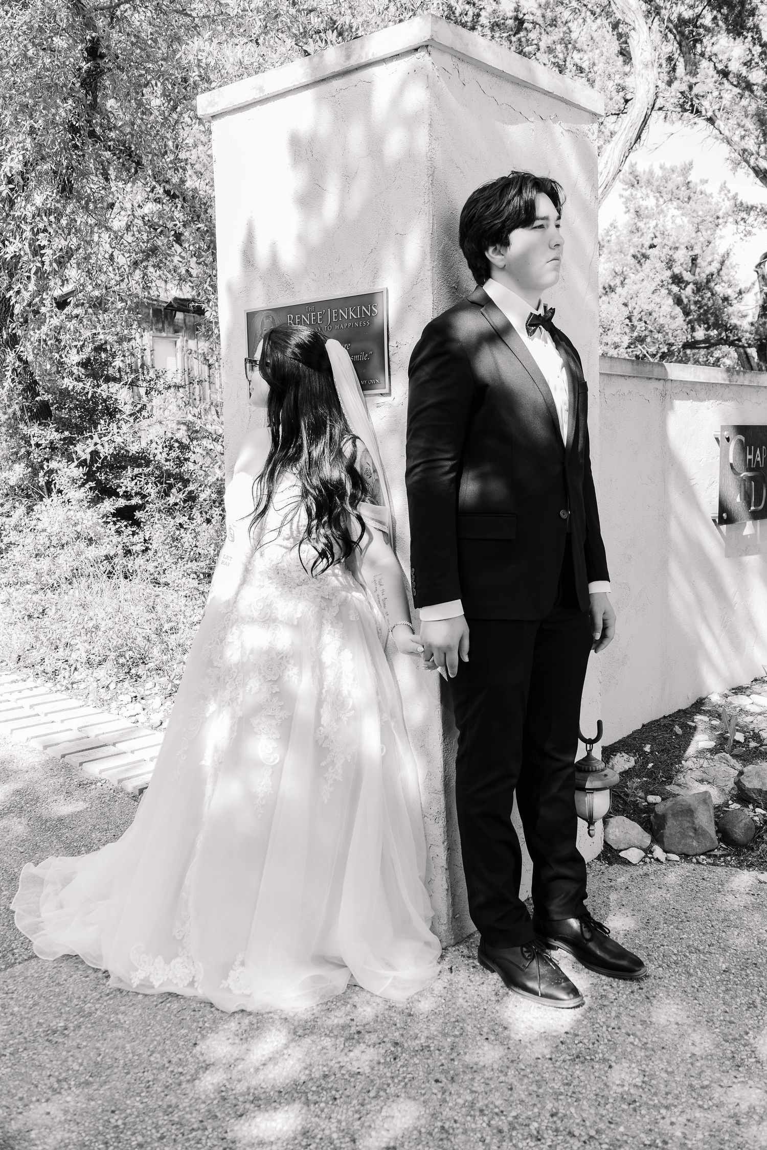 Black and white photo of a bride and groom holding hands, standing before a monument outdoors on their wedding day.