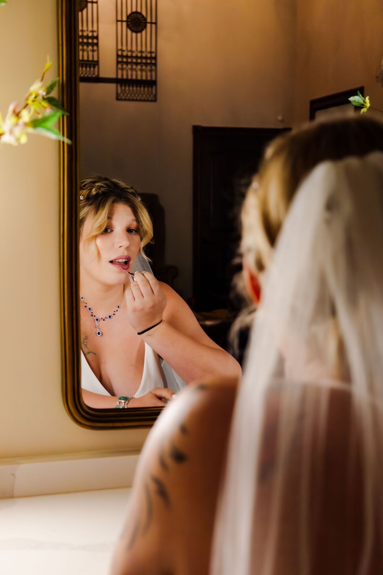 Bride applies lipstick while looking in a mirror, wearing a blue gemstone necklace on her wedding day.