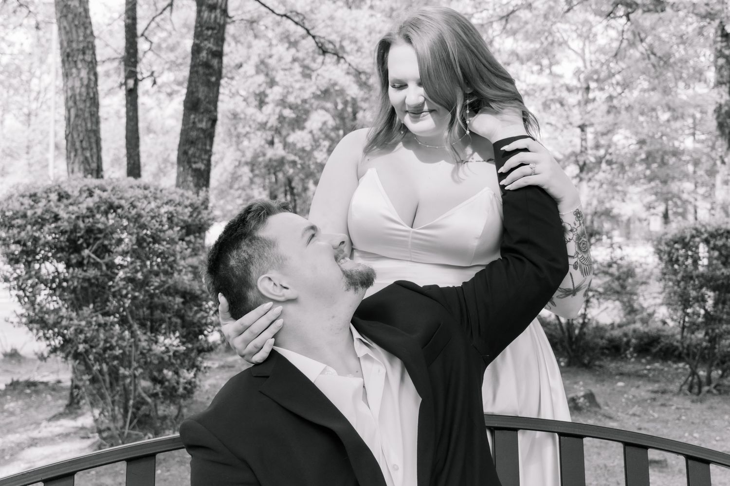 Black and white romantic photo of a couple sharing a tender moment on a wooden bridge in a park.