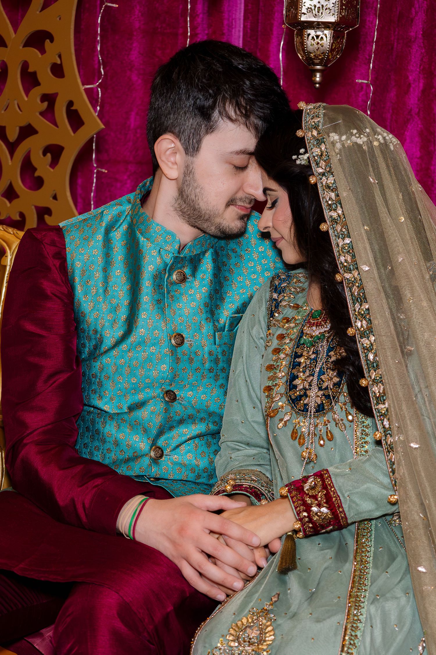 Couple in traditional South Asian wedding attire, groom in teal sherwani, bride in embroidered mint lehenga, sharing intimate moment.