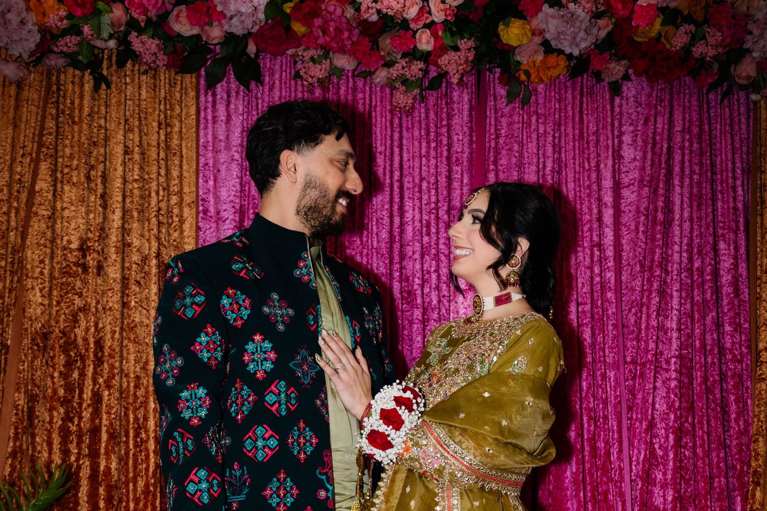 Indian couple in traditional wedding clothes gazing at each other before a pink velvet and floral backdrop.