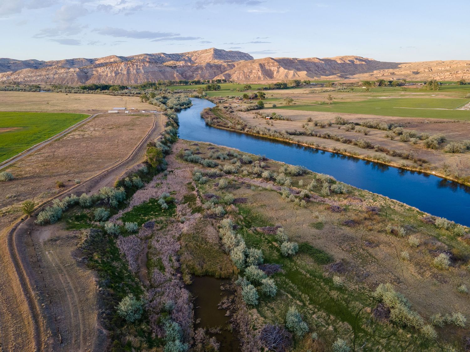 Aerial view of a winding river through a lush valley with green fields and rugged mountains at golden hour.