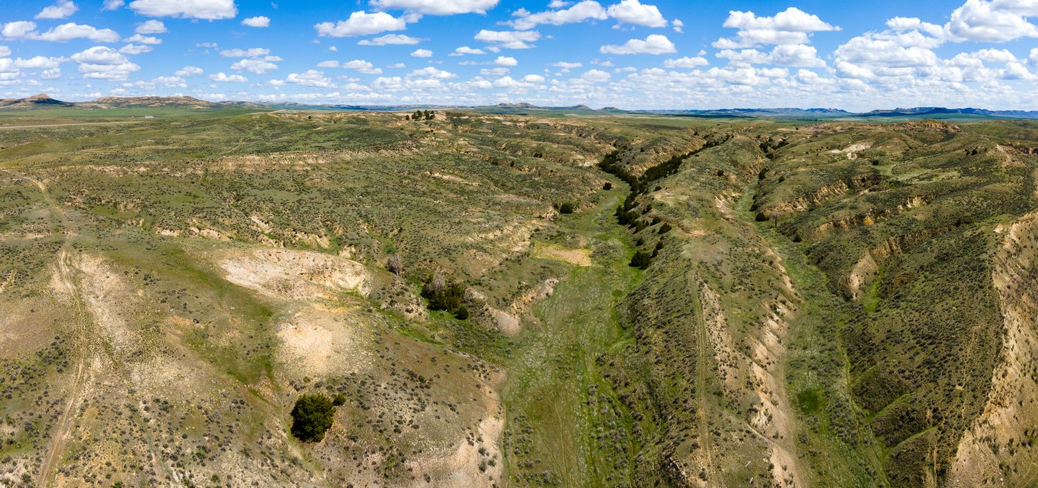 Aerial panoramic view of a rugged canyon landscape with green vegetation under a partly cloudy blue sky on a Wyoming ranch.