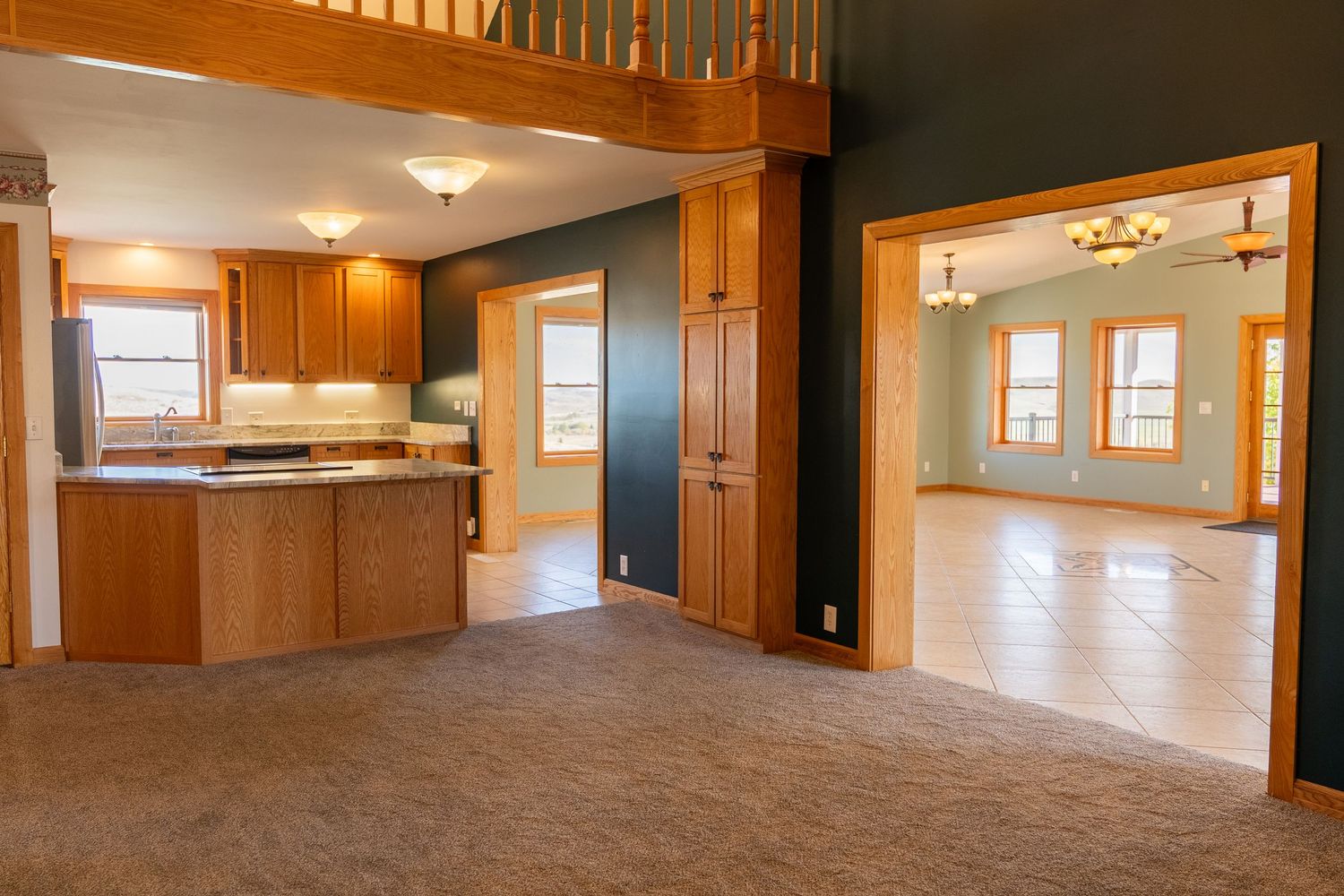 Open-concept kitchen and living area with warm wood cabinetry, carpet flooring, and bright windows in a spacious home.