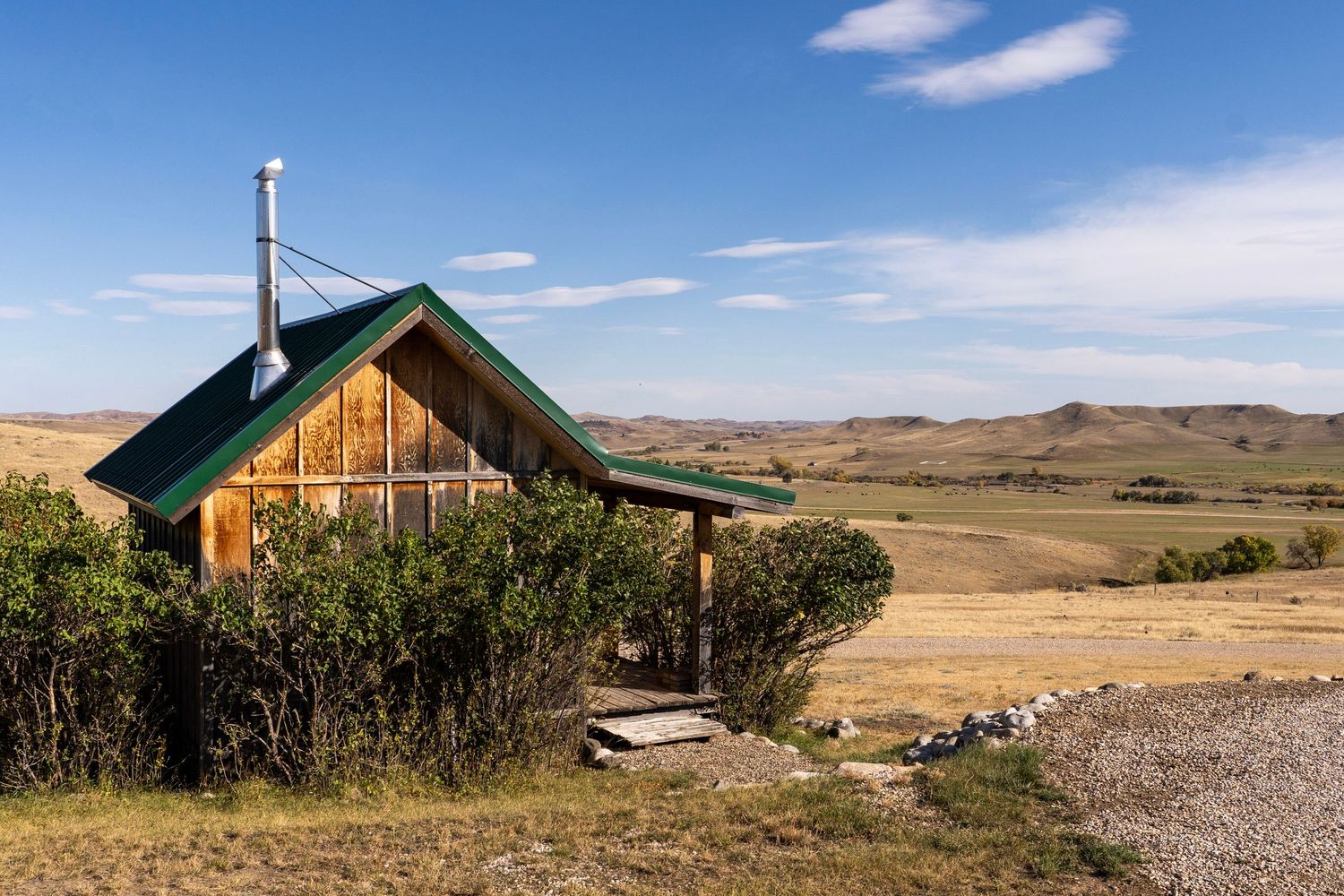 A rustic wooden cabin with a green roof sits amid shrubs overlooking vast rolling prairie hills under a blue sky.