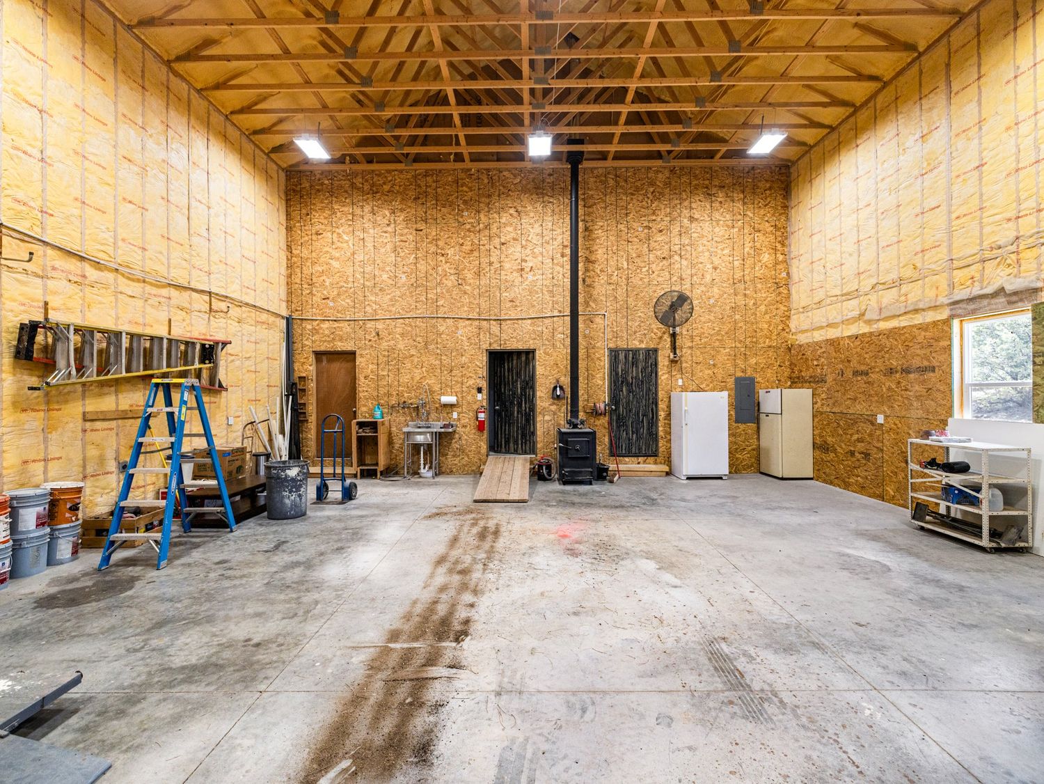Spacious wood-paneled workshop interior with high ceilings, concrete floor, blue ladder, and wood-burning stove.