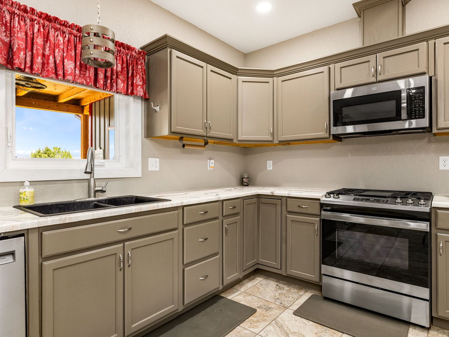 Modern kitchen with gray cabinets, stainless steel appliances, gas range, microwave, and sink under a window with red valance.