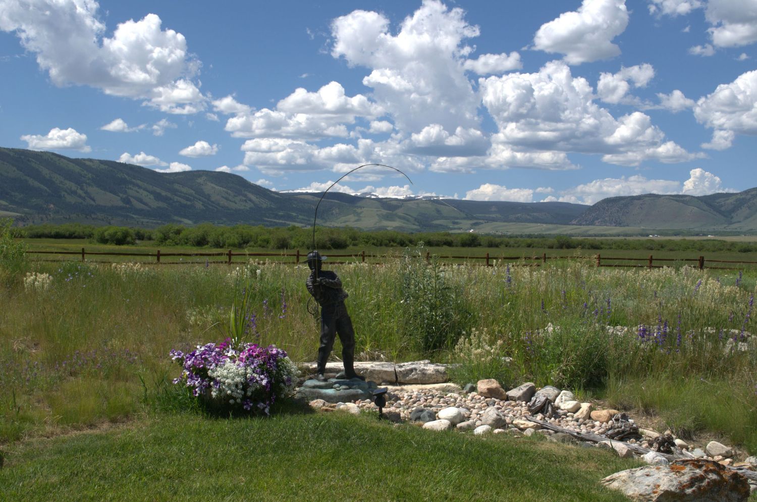 Fishing statue standing near wildflowers and a rock stream in a scenic mountain valley with green fields and cloudy blue sky.