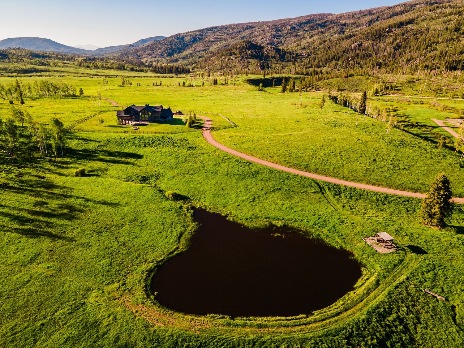 Aerial view of a lush green ranch with a heart-shaped pond, winding dirt road, red barn, and forested mountain backdrop.