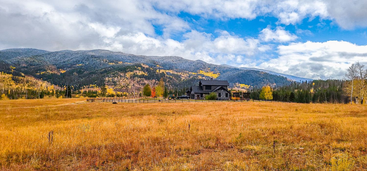 Rustic Colorado mountain lodge surrounded by golden autumn meadows with colorful fall foliage and Rocky Mountain peaks under blue skies.