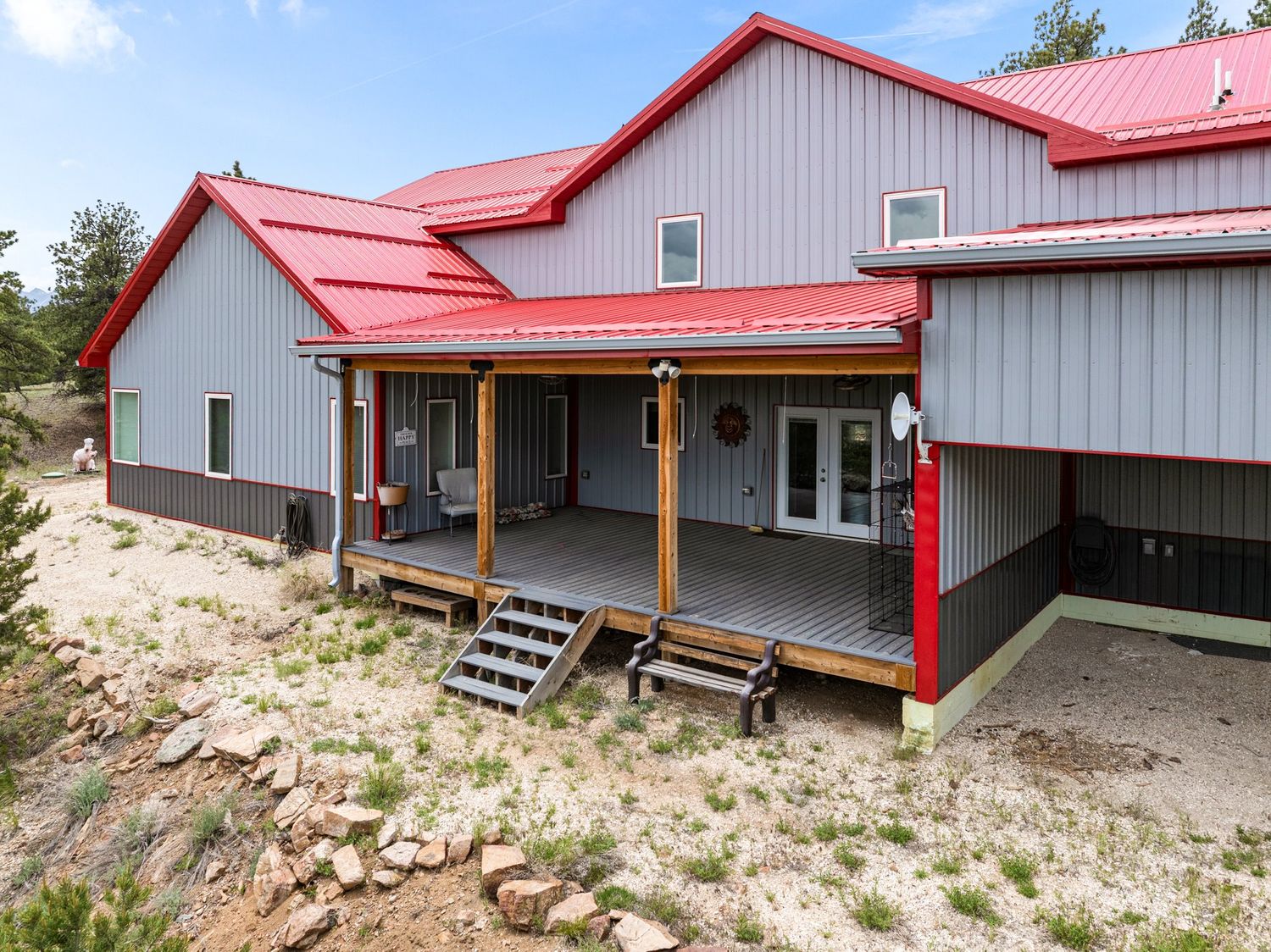 Metal barndominium with red roof and covered porch surrounded by natural Colorado landscape and pine trees.