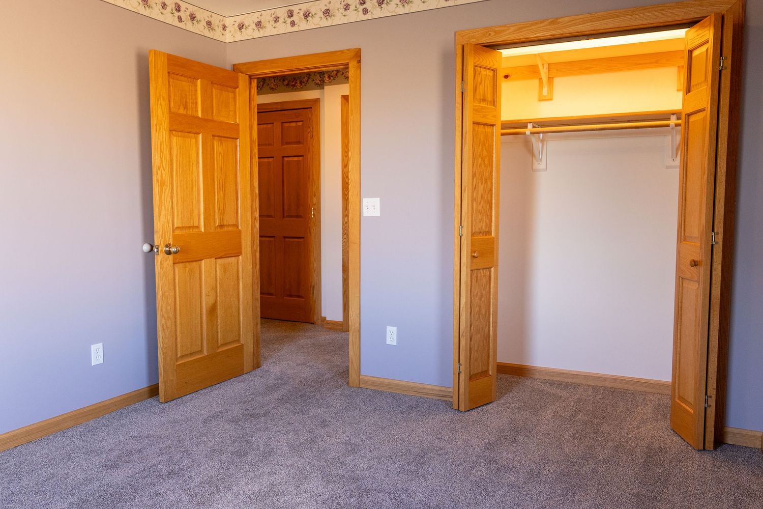 Empty bedroom with gray carpet, open closet with bifold doors, and wooden interior doors in a neutral-toned room.