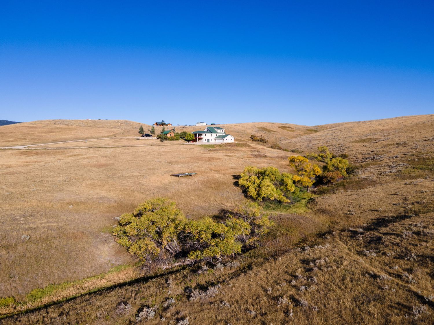 Aerial view of a remote farmhouse on golden rolling hills under a clear blue sky with sparse trees nearby.