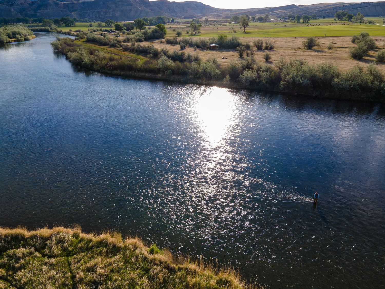 Aerial view of a serene river winding through green fields and hills, with sunlight reflecting off the calm water surface.