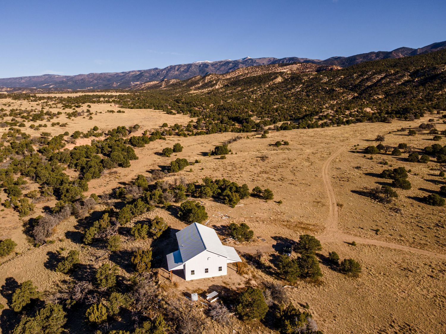 Aerial view of a white barn surrounded by juniper trees in a vast arid landscape with mountain range backdrop.