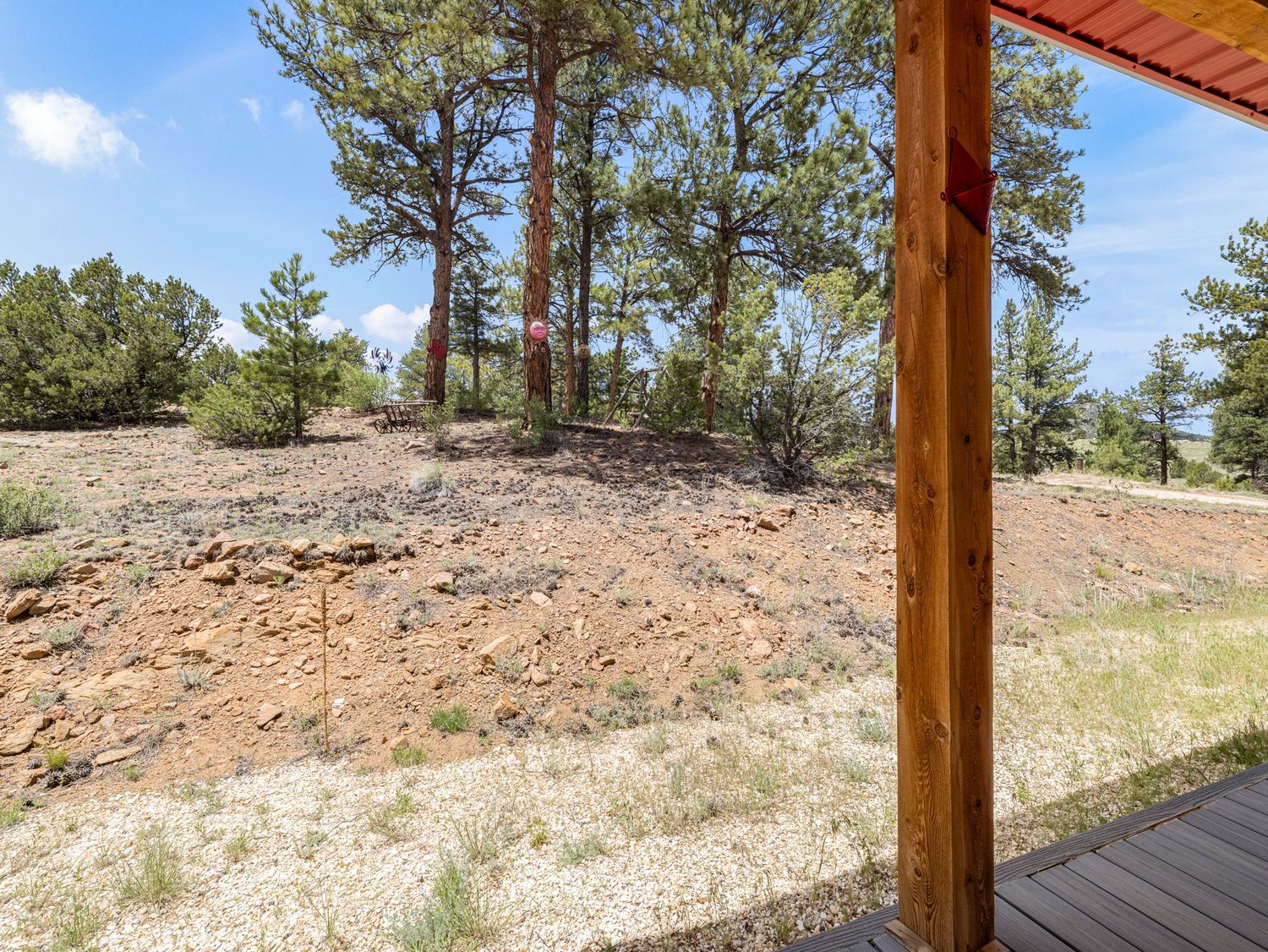 Rustic wooden deck overlooks a dry, rocky landscape with tall pine trees under a partly cloudy blue sky.