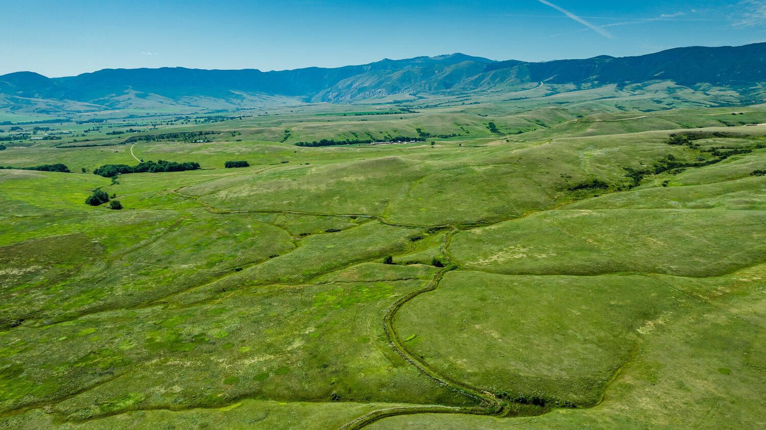 Aerial view of lush green rolling hills and valleys with mountain range in the background under a clear blue sky.