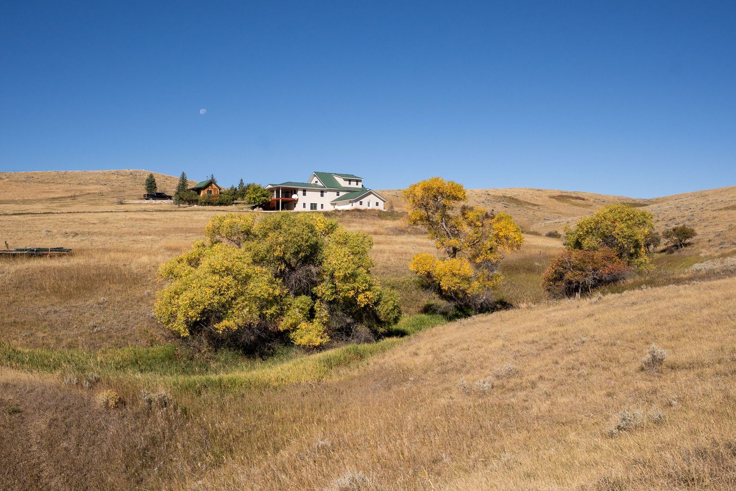 A white farmhouse nestled among golden autumn trees on rolling prairie hills under a clear blue sky.