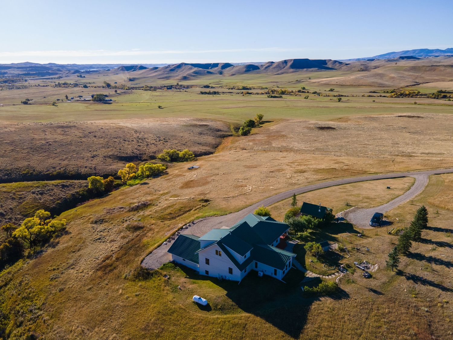 Aerial view of an isolated ranch house surrounded by vast open grasslands and rolling hills under a clear blue sky.