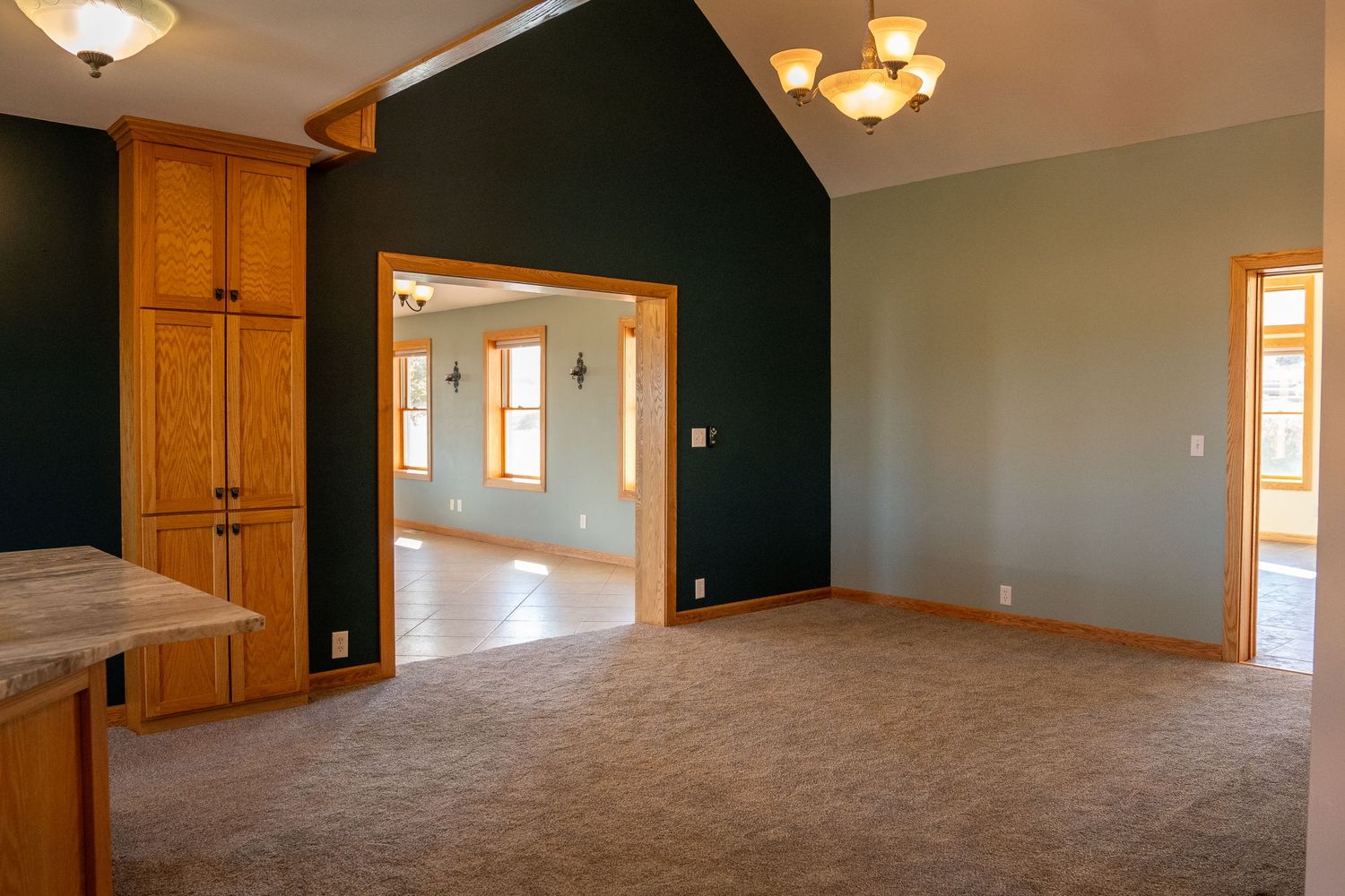 Spacious open-concept living area with vaulted ceilings, dark green accent walls, beige carpet, and warm wood trim details.
