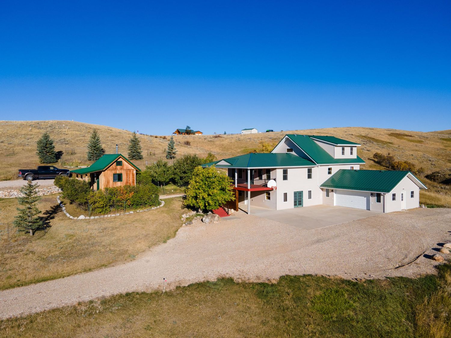 Aerial view of a rural property with green-roofed white farmhouse, detached garage, and log cabin set among rolling hills.